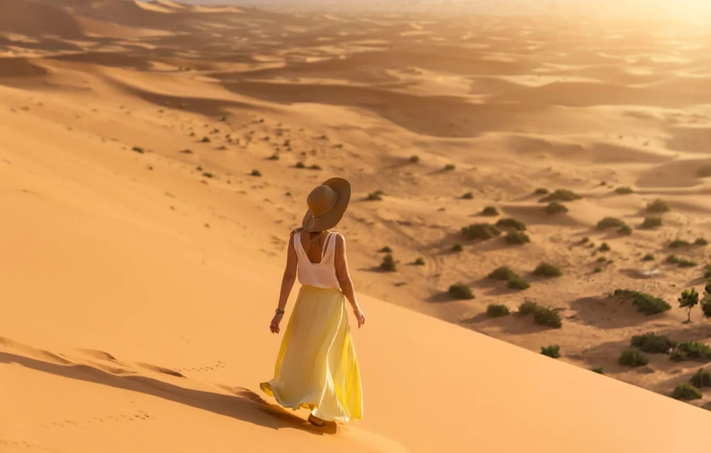 A young woman in a long yellow skirt, white shirt and straw hat is watching the dawn from the top of the sand dune of the Merzouga desert. Dawn in the Sahara desert.